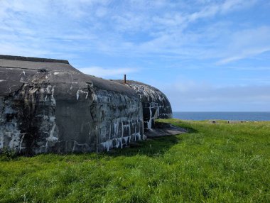 Yıpranmış beton sığınak yeşil çimenli tepenin üzerinde duruyor açık gökyüzünün altında açık bulutlarla mavi okyanusa bakan