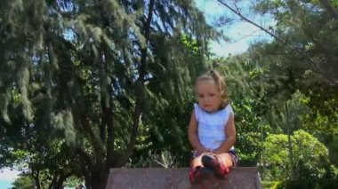 beautiful little baby sitting on a granite pedestal and looks around. she smiles and straightens her hair. in the background trees are.