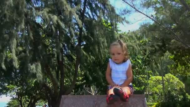 Beau petit bébé assis sur un piédestal de granit et regarde autour. elle sourit et redresse ses cheveux. dans les arbres de fond sont . — Video