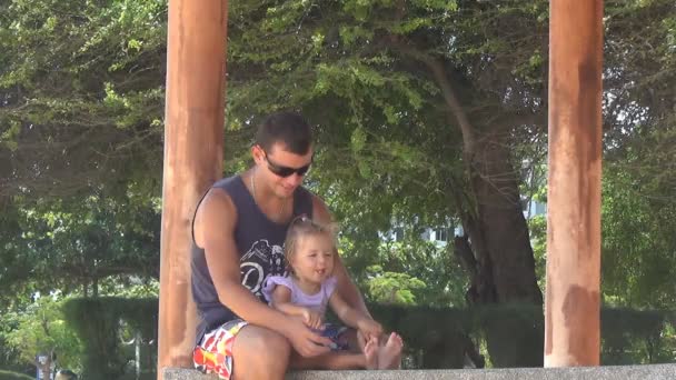 Papa und Baby spielen im Pavillon am Meer. Mädchen stampft Füße. Papa behält die Beine der Tochter. sie lachen. — Stockvideo