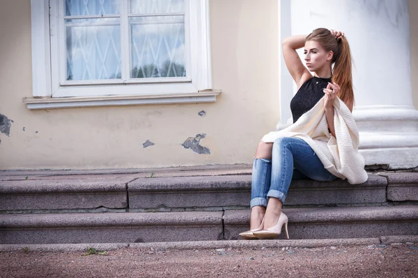 Красивая девушка сидит на каменной лестнице. Она выпрямляет джинсы. мода — стоковое фото