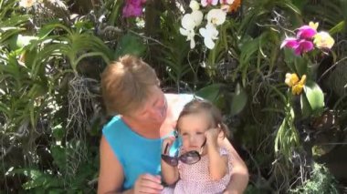 mavi elbiseli bir kadın kucağına oturan ve ağaçları ve güzel Orkide çok renkli arka plan üzerinde güneş gözlüğü ile oynarken, bebek. çocuk alır ve sonra koyar güneş gözlüğü