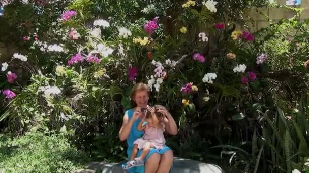 Bébé assis sur les genoux d'une femme en robe bleue et jouant avec des lunettes de soleil sur le fond d'arbres et de belles orchidées multicolores — Video