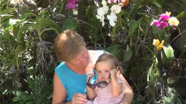 Bambino seduto sulle ginocchia di una donna in abito blu e giocare con gli occhiali da sole sullo sfondo di alberi e belle orchidee multicolori. il bambino prende, e poi mette occhiali da sole — Video Stock