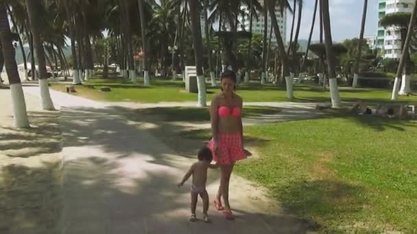 Νέοι όμορφη γυναίκα σε ένα ροζ κοστούμι λουσίματος με ένα μωρό περπάτημα στην προκυμαία στο φόντο της ένα τροπικό πάρκο και κουνώντας το χέρι του στην κάμερα — Αρχείο Βίντεο
