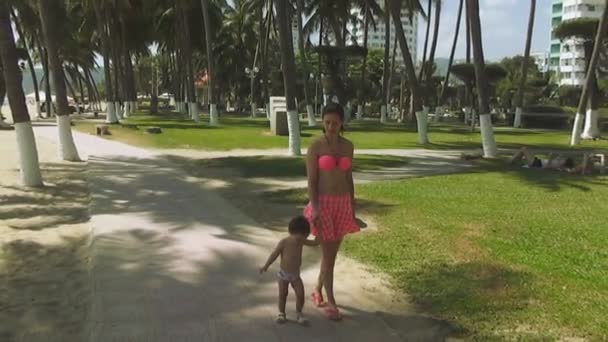 Jovem mulher bonita em um maiô rosa com um bebê andando na orla marítima no fundo de um parque tropical e acenando com a mão para a câmera. Movimento lento — Vídeo de Stock