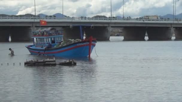 Vietnamesisches blaues Boot vor Anker auf dem Fluss. Fischer werfen Fischernetze ins Boot. Im Hintergrund eine Brücke mit fahrenden Autos und Fahrrädern — Stockvideo