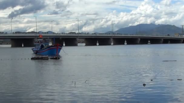 Vietnamesisches blaues Boot vor Anker auf dem Fluss. Fischer werfen Fischernetze ins Boot. Im Hintergrund eine Brücke mit fahrenden Autos und Fahrrädern — Stockvideo