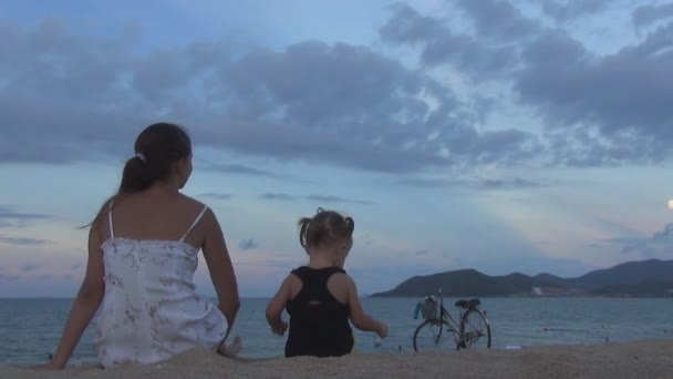 Belle fille aux cheveux longs avec un bébé assis le dos à l'appareil photo sur la plage de sable fin. ils regardent le ciel bleu et une grande lune. Pleine lune . — Video