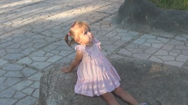 Beau bébé assis sur un grand rocher. Elle porte une robe. Elle regarde le ciel et sur les côtés. bébé a vu quelque chose, elle est surprise et pointe vers le côté . — Video