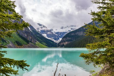Louise Gölü ağaçlar tarafından çerçevelenmiş dağlara ve bulutlu gökyüzüne bakan yansımalı Kanada