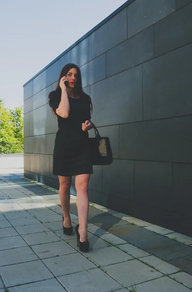 Biznesowe dziewczyna w czarnej sukni, w pobliżu ściany, rozmawiając przez telefon — Zdjęcie stockowe