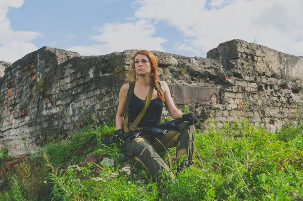 Vacker rödhårig flicka i militär uniform sitter på en förstörd byggnad med en pistol i handen — Stockfoto