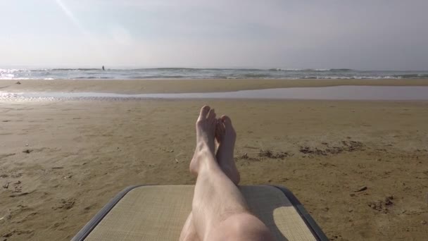 Die Beine der Männer auf einer Chaiselongue vor dem Hintergrund eines Sandstrandes und des Meeres — Stockvideo