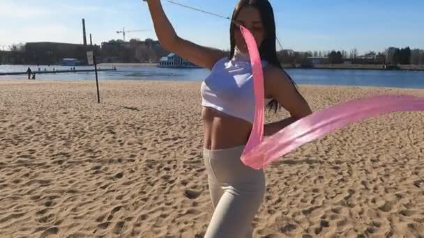 O em uma menina de manhã ensolarada em uma praia arenosa faz exercícios com uma fita de ginástica — Vídeo de Stock