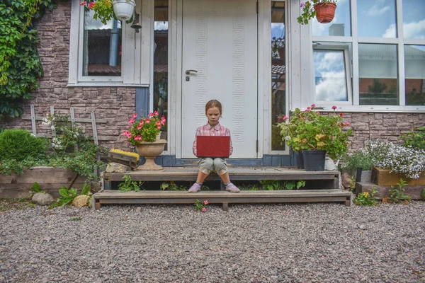 a girl child with a laptop is sitting on the porch of the house during a lesson on a remote photo without a filter