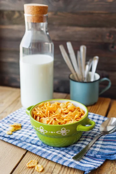 Tasty corn flakes in green bowl with bottle of milk. Rustic wooden background with plaid blue napkin. Healthy crispy breakfast snack.