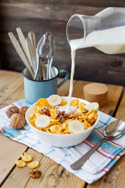 La leche de una botella fluye en el tazón con sabrosos copos de maíz con nueces y plátano. Fondo rústico de madera con servilleta a cuadros. Snack crujiente saludable para el desayuno . — Foto de Stock