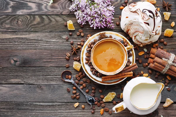 Rustic wooden background with cup of coffee, milk, meringue, cinnamon and lilac flowers. White vintage dinnerware and spoon. Breakfast at summer morning. Top view, place for text. — Stock Photo, Image