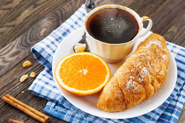 Desayuno continental - taza de café caliente, croissant y naranja. Sabrosa comida en servilleta azul a cuadros, fondo rústico de madera . —  Fotos de Stock