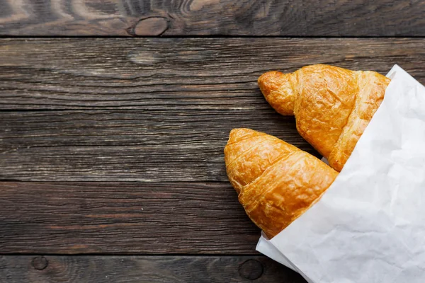 Tasty croissants in white paper bag. Rustic wooden background with sweet dessert. Top view. — Stock Photo, Image