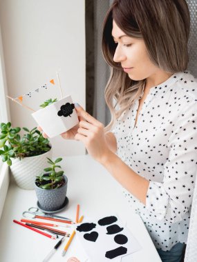 Gülümseyen kadın Cadılar Bayramı için el yapımı süslemeler sergiliyor. DIY bayrakları ve Boo! Çiçek saksısına yapışkan bitkilerle yapıştırılmış. Evde sosyal açıdan uzak cadılar bayramı.