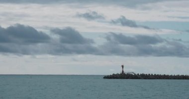 Martılar ve karabataklar dalgakıranların üzerinde otururlar. Bulutlu arka planda deniz feneri. Soçi Limanı, Rusya.