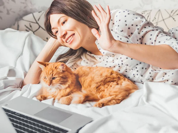 Cute ginger cat and woman are lying in bed. Woman is greeting somebody via online video translation or video call. Online communication. Morning bedtime with fluffy pet.