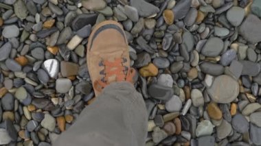 Kahverengi spor ayakkabılı adam çakıl taşları üzerinde yürür. Ağır çekim. Kayalık plajda spor ayakkabılarla üst manzara.