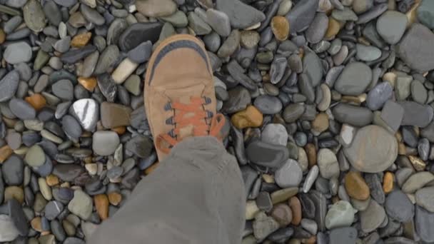 茶色のスポーツシューズの男が小石を歩く。スローモーション。岩の多いビーチでのスニーカーの上からの眺め. — ストック動画