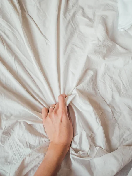 Woman\'s hand on background of crumpled bed sheet. Creased unmade bed. Woman squeezes white linen. Cozy imperfect house. Non-ideal cozy home.