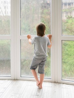 Küçük çocuk ıslak panoramik pencereden yağmur damlalarıyla dışarı bakıyor. Coronavirus COVID19 karantinası. Yağmurlu yaz havası. Çocuk evde yalnız kalıyor. Çocuksuz yalnızlık ve hüzün..