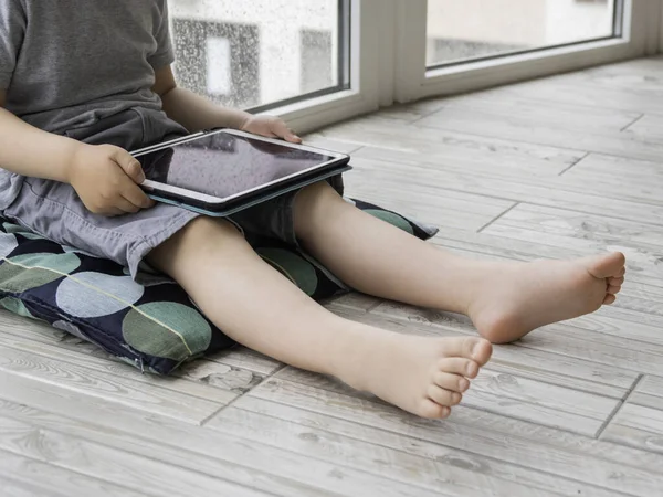 Curious boy watch cartoons on digital tablet. Kid sits on floor and uses electronic device. Indoor leisure for children while it\'s raining outside.