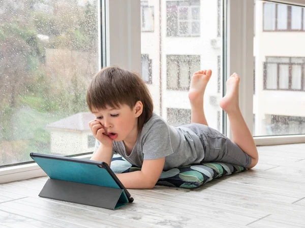 Curious boy watch cartoons on digital tablet. Kid lies on floor and uses electronic device. Indoor leisure for children while it\'s raining outside.
