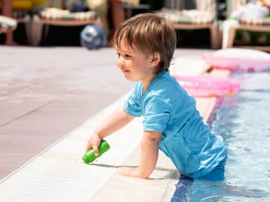 Yüzme havuzunda yıkanan tatlı bebek. Parlak mavi mayo giyen küçük çocuk havuzda oynuyor. Otelde tatil. Plaj aksesuarları - güneş şezlongları, lastik yüzük, oyuncaklar.