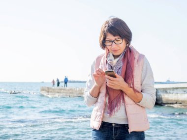 Gülümseyen kadın akıllı telefondan birine mesaj atıyor. Deniz kenarında aktif bir eğlence. Okyanus kıyısında tatil. Kadının arkasında mutlu bir gülümsemeyle turkuaz su..