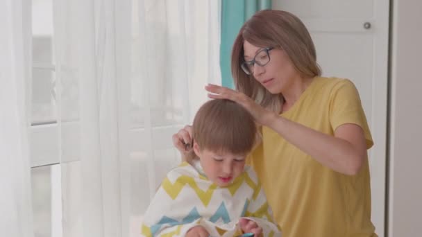 Mor själv klipper sina söner hår hemma. Ny normal. Pojken sitter täckt med tyg och leker med sax. Livsstil, egenvård. — Stockvideo