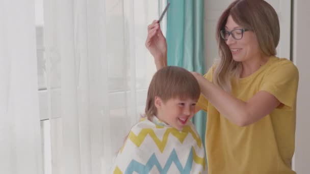 Mor selv klipper hendes sønner hår derhjemme. Ny normal. Dreng sidder dækket med klud og leger med saks. Livsstil, egenomsorg. – Stock-video