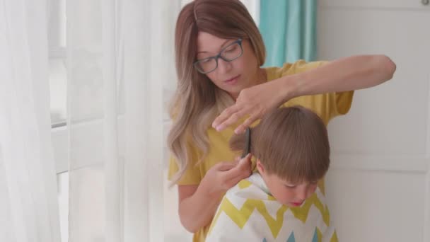 Sama matka obcina synom włosy w domu. Nowa normalność. Chłopiec siedzi pokryty tkaniną i bawi się nożyczkami. Styl życia, Samoopieka. — Wideo stockowe