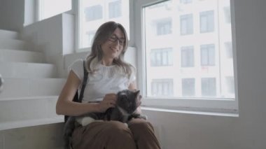 Güzel bir kadın, apartmanın beyaz merdivenlerinde otururken renkli sokak kedisini okşar. Evsiz kedi zevkle mırıldanıyor.