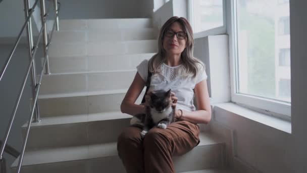 Smuk kvinde strøg farverige omstrejfende kat, mens du sidder på hvide trin af trappe af lejlighedskompleks. Hjemløse kat spinder med glæde. – Stock-video