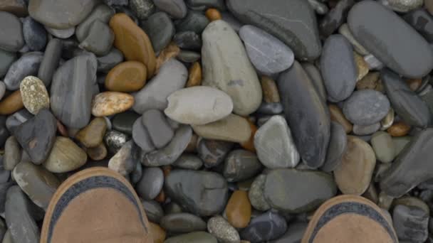 Hombre en zapatos deportivos marrones camina sobre guijarros. En cámara lenta. Vista superior de los pies en zapatillas de deporte en la playa rocosa. — Vídeo de stock