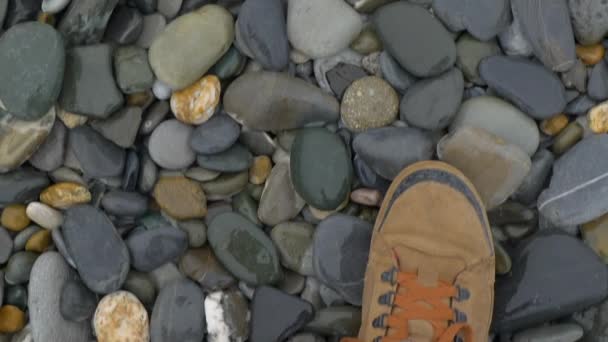 Чоловік у коричневому спортивному взутті ходить на гальку. Повільний рух. Вид зверху на ноги в кросівках на кам'янистому пляжі . — стокове відео