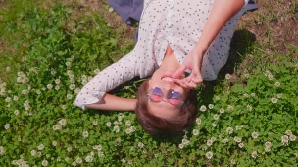 Top view on woman with colorful sunglasses lying on lawn in urban park and enjoys flowers. Summer heat in town. — Stock Video