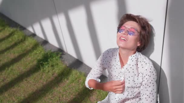 Žena s barevnými slunečními brýlemi sedí na trávníku v městském parku a těší se jasnému slunečnímu svitu. Letní vedro ve městě. — Stock video