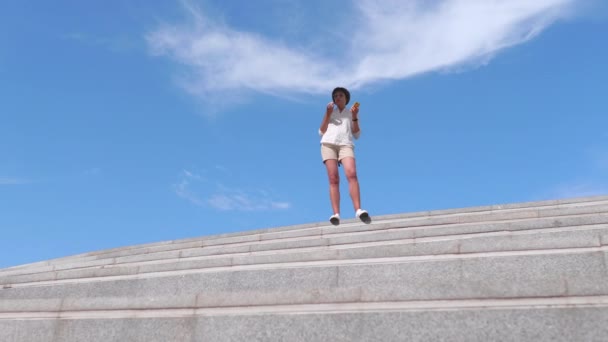 여성은 도시 공원의 계단 위에 서 있습니다. 암컷은 맑고 푸른 하늘 배경에서 사과를 먹는다. 여름 경치. — 비디오