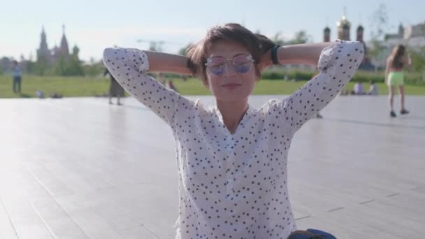 Mujer con gafas de sol de colores se sienta en escena en el parque urbano y disfruta de la luz del sol brillante. Calor de verano en la ciudad. — Vídeos de Stock