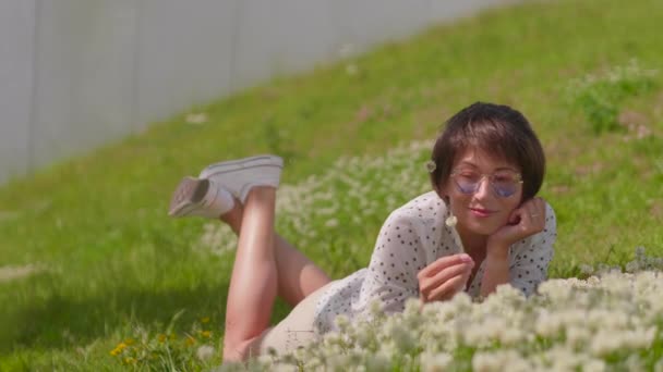 Renkli güneş gözlüklü kadın şehir parkında çimlere uzanıyor ve çiçeklerden hoşlanıyor. Kasabada yaz sıcağı. — Stok video