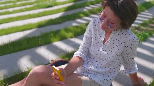 Woman with colorful sunglasses sits on lawn in urban park and makes selfie on smartphone. Summer heat in town. — Stock Video