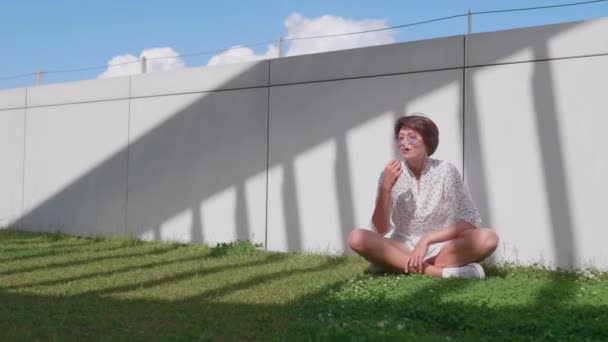 Femme avec des lunettes de soleil colorées se trouve sur la pelouse dans le parc urbain et bénéficie d'un soleil éclatant. Chaleur estivale en ville. — Video